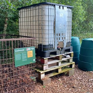 A metal cage with an open top that has piles of leaves next to it. Next to the cage are 3 wooden pallets stacked on top of each other and a Green Johanna compost bin