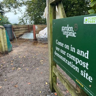A gate, opened and leading to a compost area. The gate has a dark green sign on, the sign reads "come on in and see our compost demonstration site"